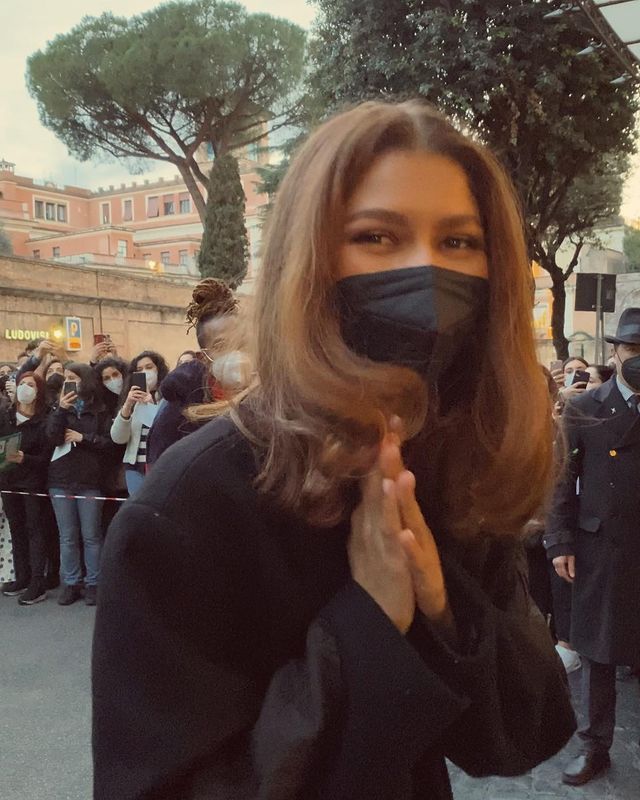 a woman wearing a black face mask standing in front of a crowd with her hands clasped