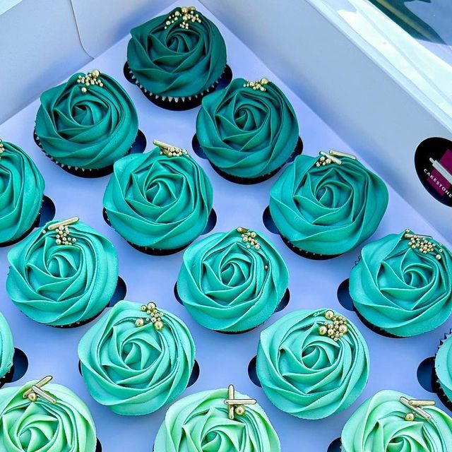 blue cupcakes with green frosting and gold decorations in a white box on a table