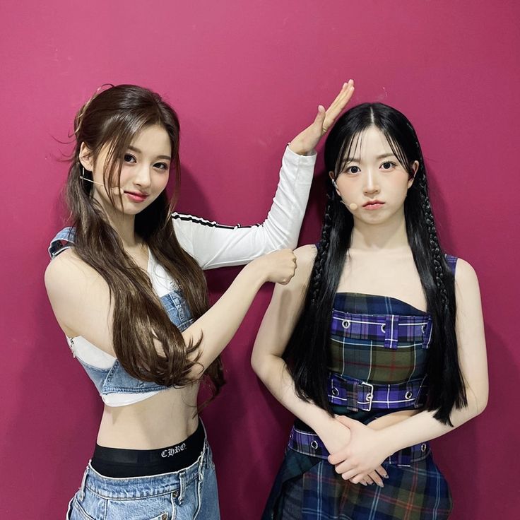 two young women standing next to each other in front of a pink wall with one pointing at the camera