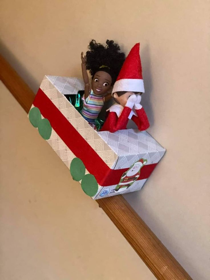 an elf is sitting in a gift box on the stair rail with her hand up to her head