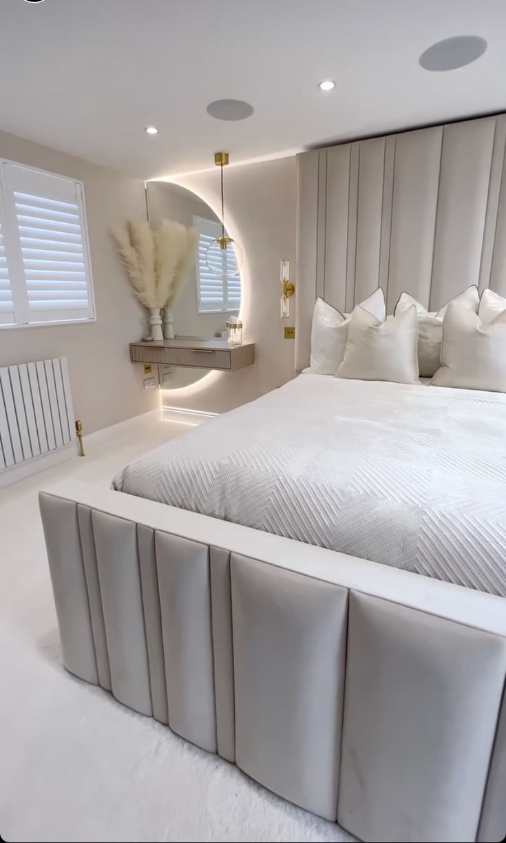 a large white bed sitting in a bedroom next to a tall wall mounted mirror with lights on it