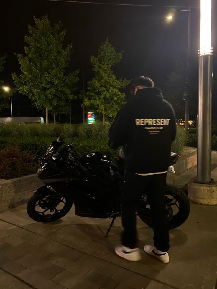 a man standing next to a parked motorcycle on a sidewalk at night with street lights in the background