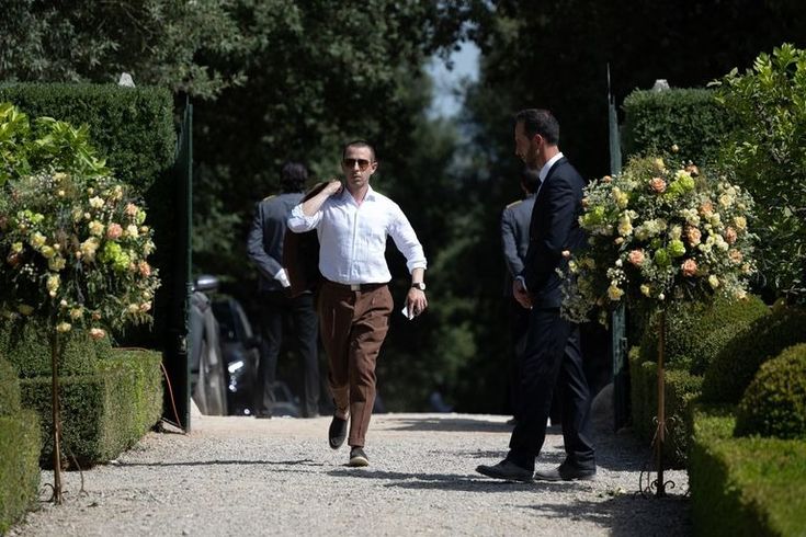 two men walking down a road next to bushes and flowers on either side of them