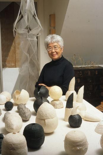 an older woman sitting at a table surrounded by clay sculptures