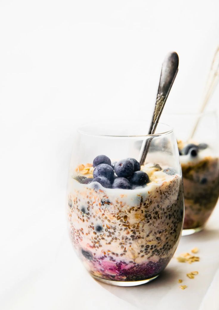 two glasses filled with cereal and blueberries on top of a white tablecloth next to silver spoons