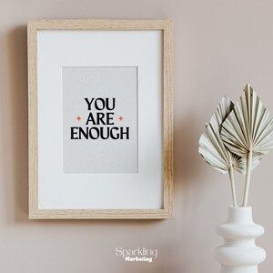 a white vase sitting next to a wall with a framed print that says you are enough