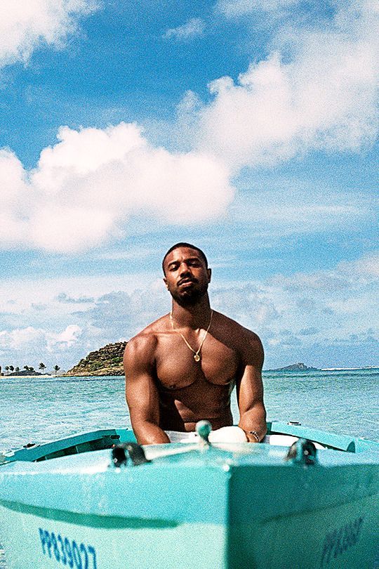 a shirtless man sitting on top of a blue boat in the ocean next to an island