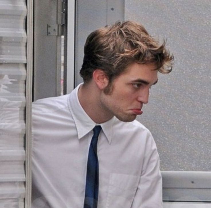 a man wearing a white shirt and blue tie sitting in front of a window with his hand on his chin