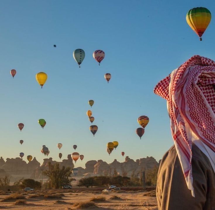 a man standing in the desert looking at hot air balloons flying over him and his head covered with a scarf