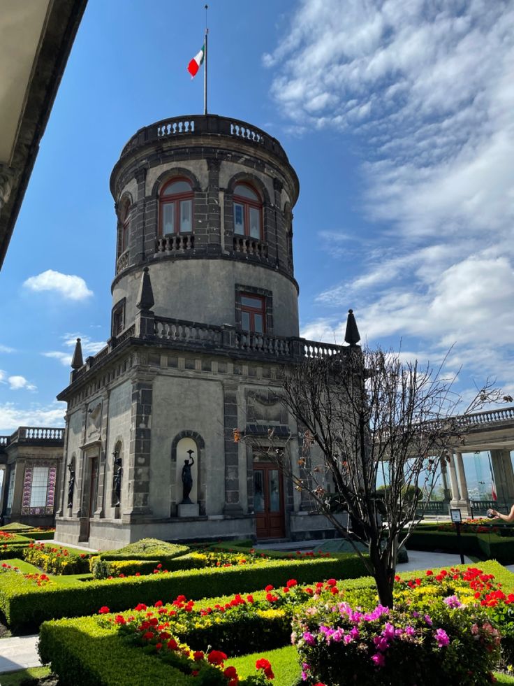 an old building with many flowers in front of it and a flag on the top