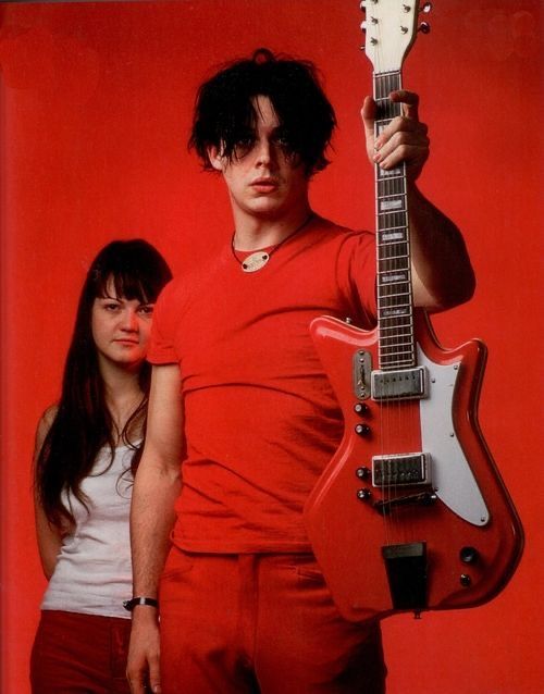 a man holding a guitar and standing next to a woman in front of a red background