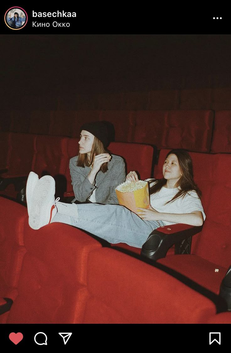 two young women sitting in the middle of a movie theater, eating popcorn and drinking soda