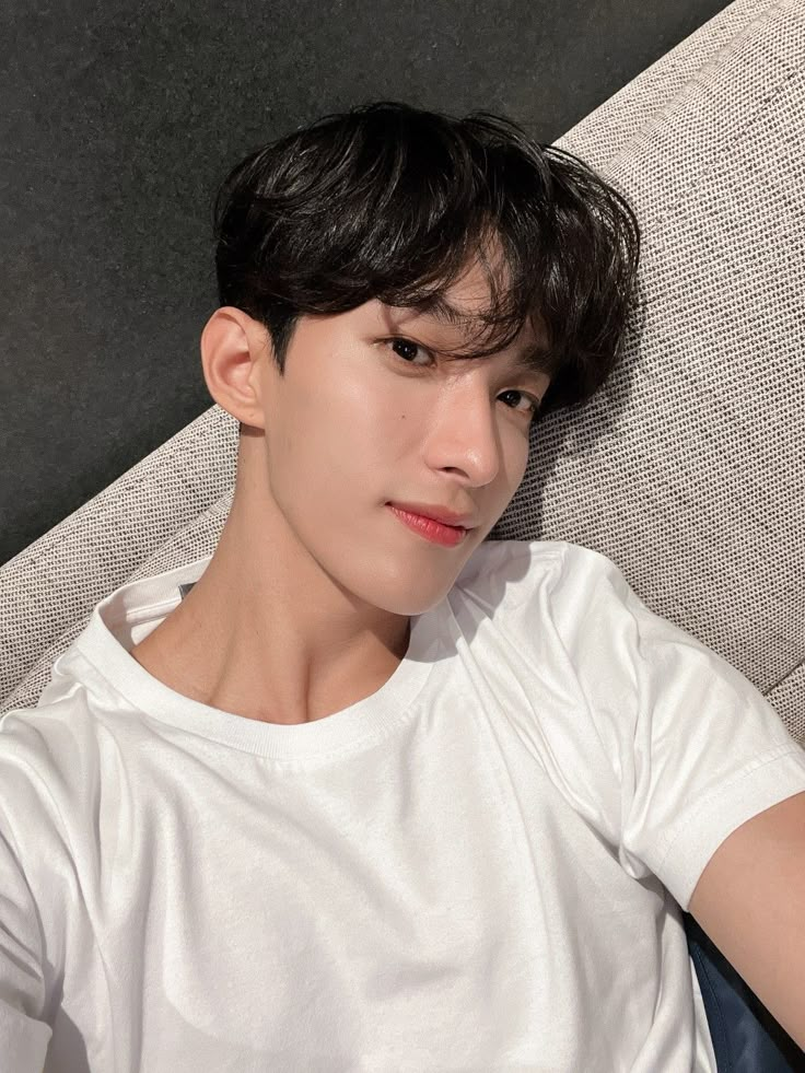 a young man sitting on top of a couch next to a white wall and wearing a white t - shirt