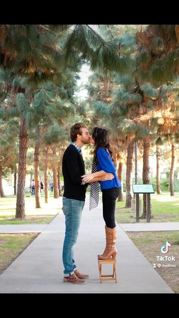 a man and woman kissing in front of pine trees on a path between two benches