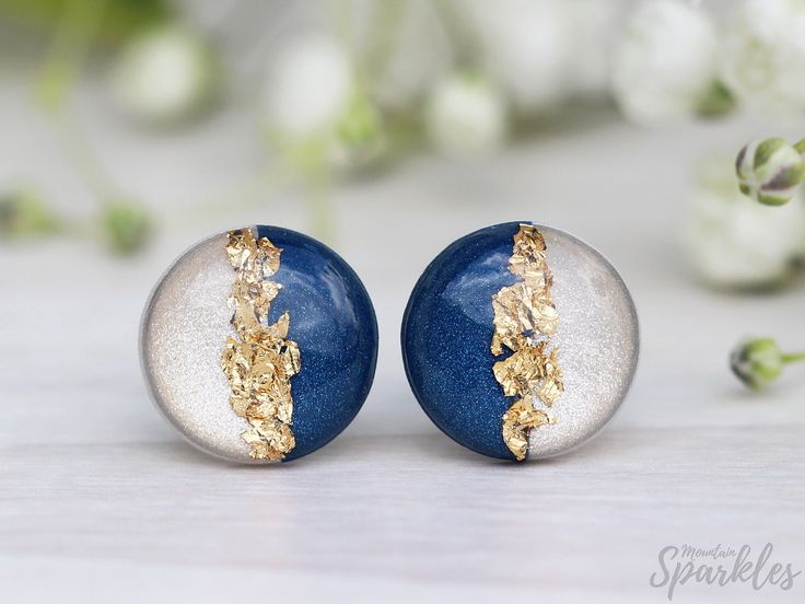 two blue and silver earrings with gold leaf detail on them sitting on a white surface