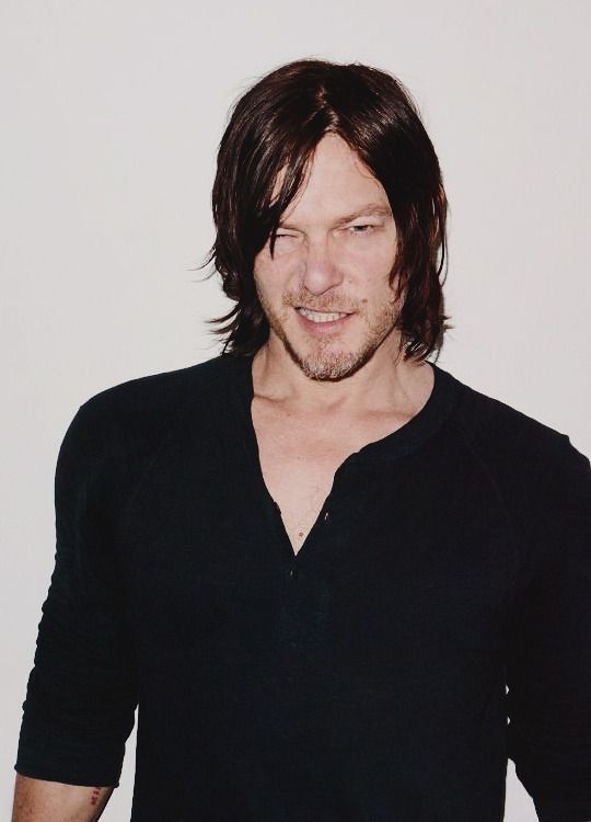 a man with long hair wearing a black shirt and looking at the camera while standing in front of a white wall