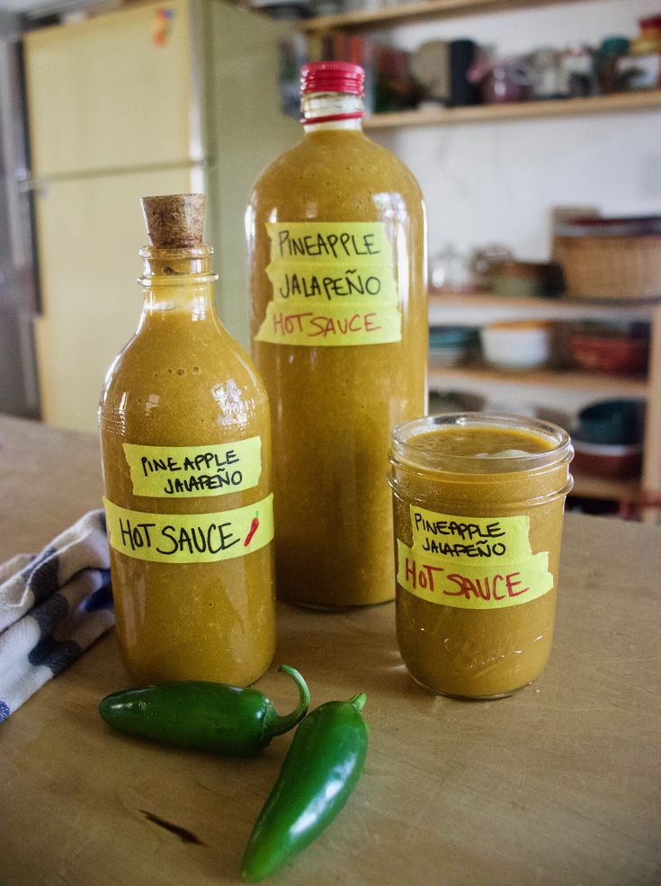 two bottles of hot sauce sitting on top of a wooden table next to green peppers