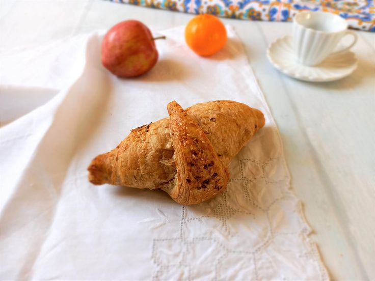 an apple and croissant sitting on a napkin next to some oranges, coffee cup and saucer