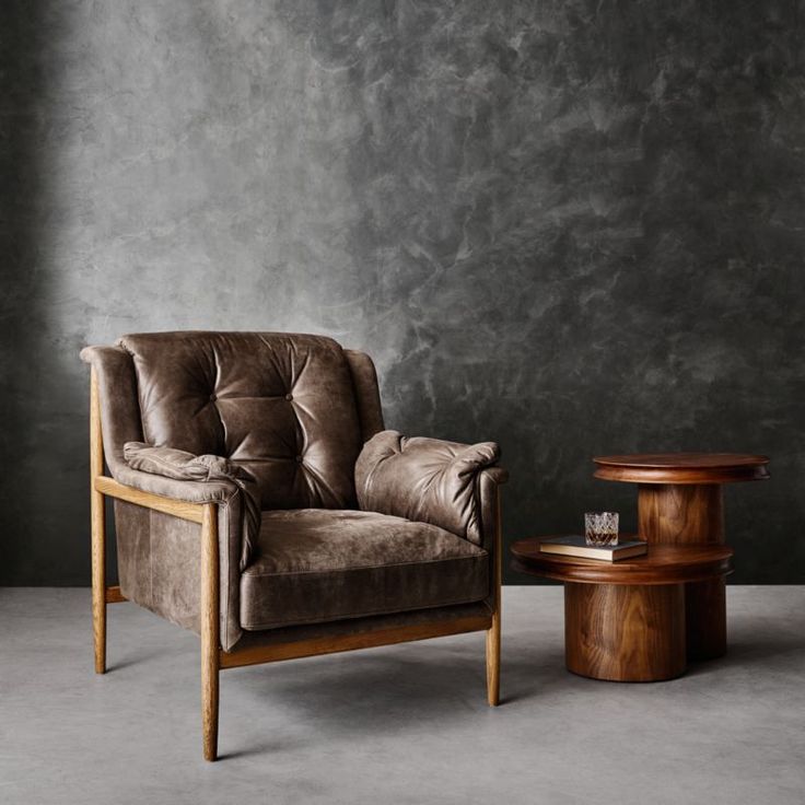 a brown chair sitting next to a wooden table on top of a cement floor with a gray wall behind it