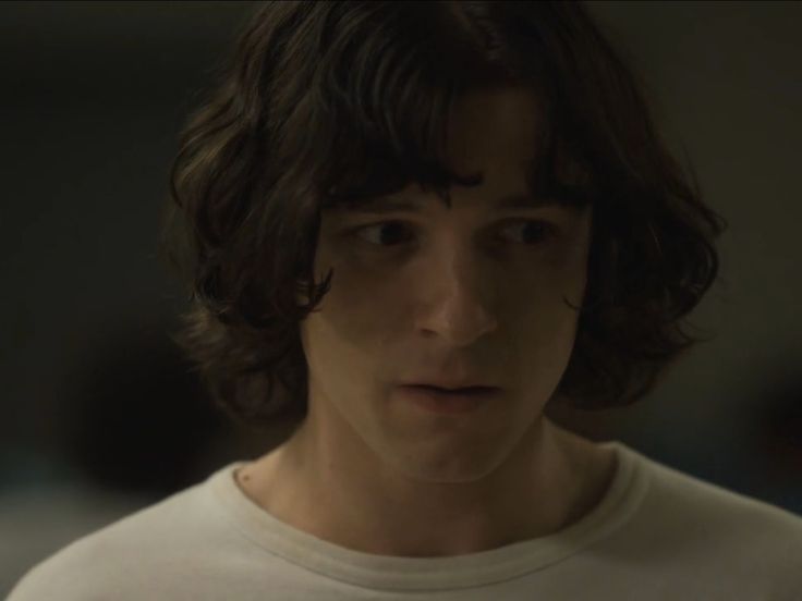 a young man with long hair wearing a white t - shirt looking at the camera