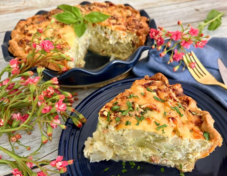 a slice of quiche on a blue plate next to a fork and pink flowers