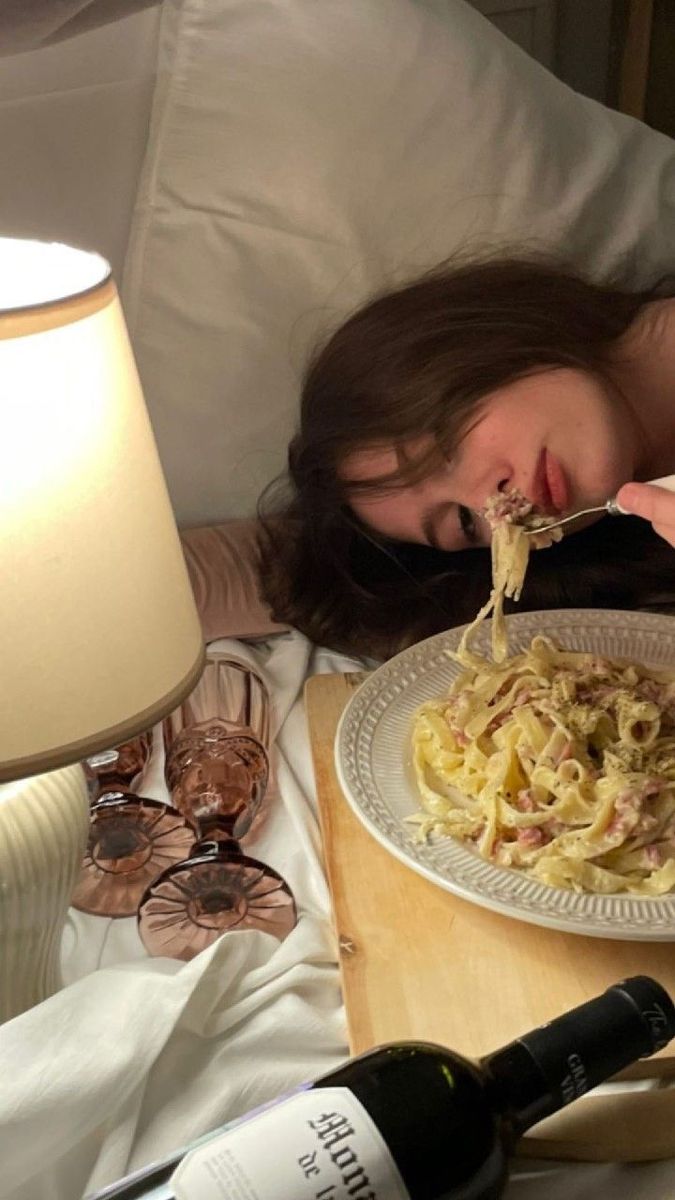 a woman laying in bed eating spaghetti with a bottle of wine next to her on the side