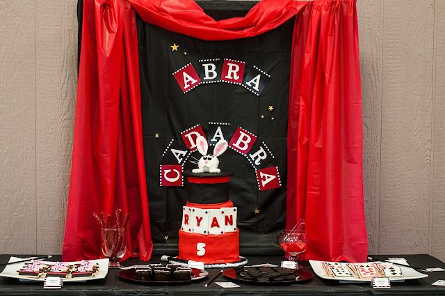 a table topped with cakes and desserts under a red drape covered backdrop that says abra