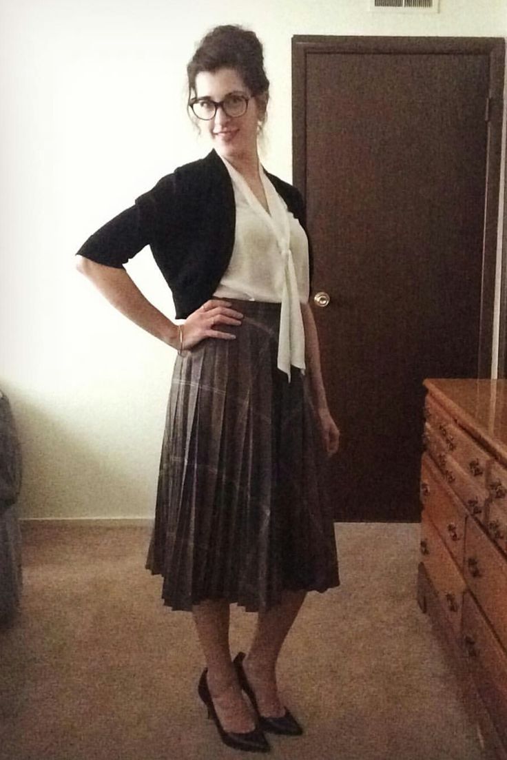 a woman standing in front of a wooden dresser wearing glasses and a white shirt with a black cardigan