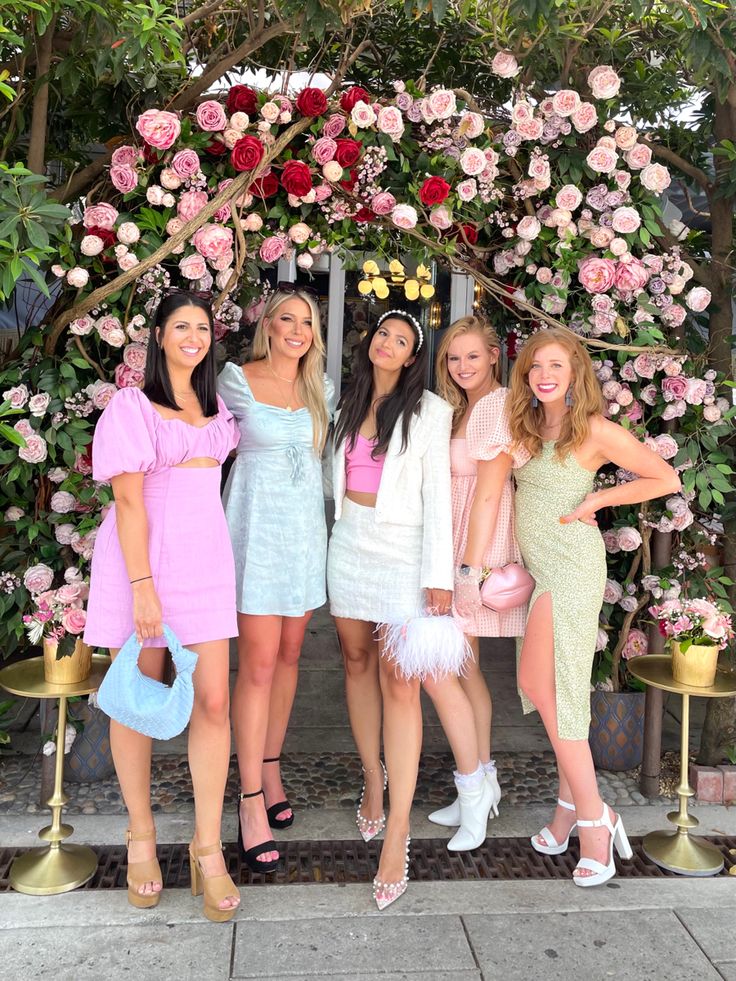 four women posing for a photo in front of a flower covered archway with pink and white flowers