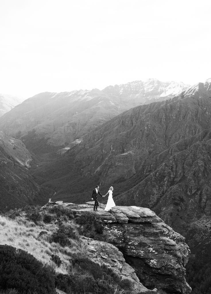 a bride and groom standing on top of a mountain looking out at the valley below