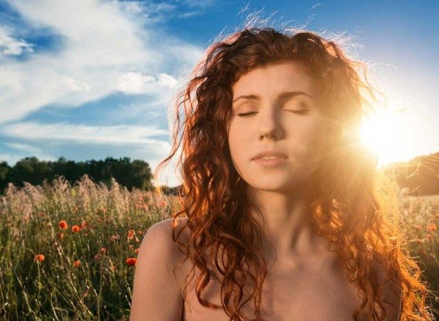 a woman with long red hair standing in a field at sundown, her eyes closed