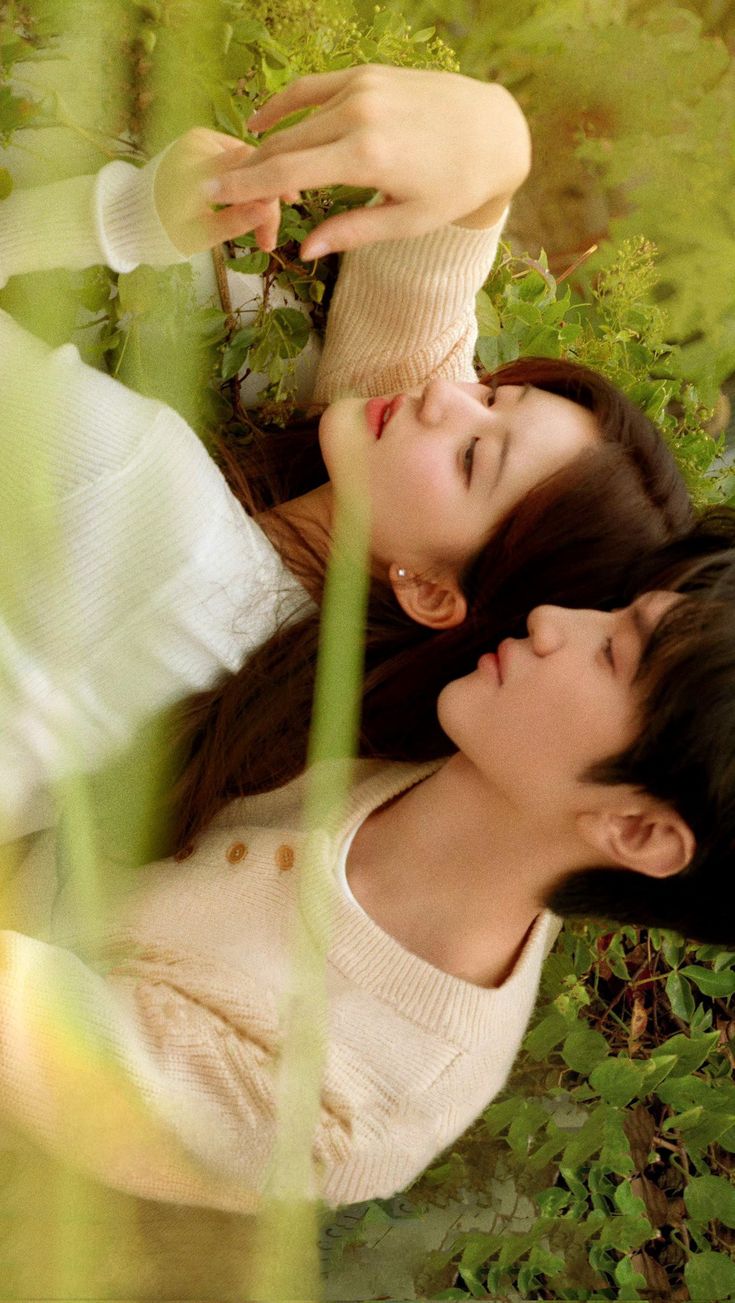 two young women laying on the ground in front of green plants, one holding her head