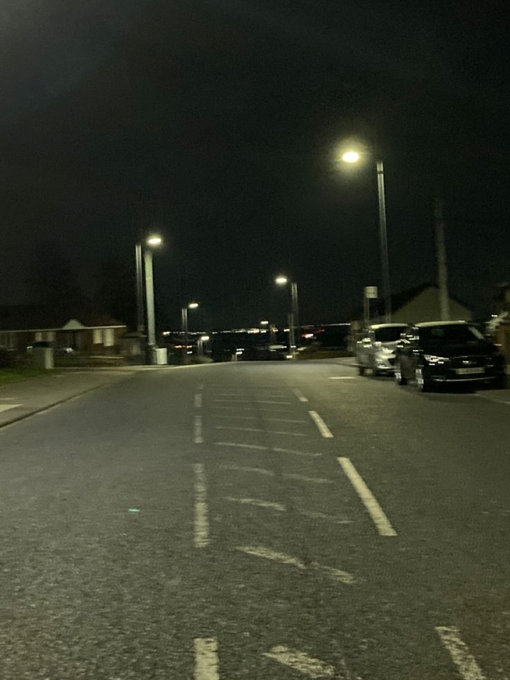 an empty street at night with cars parked on the side and lights in the background