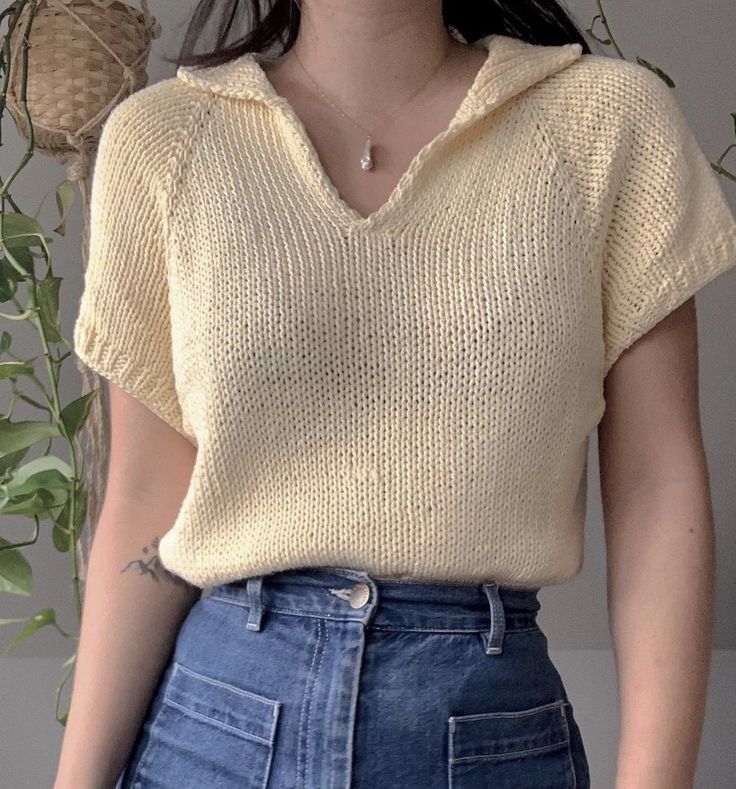 a woman wearing a yellow knitted sweater and blue denim skirt standing in front of a potted plant