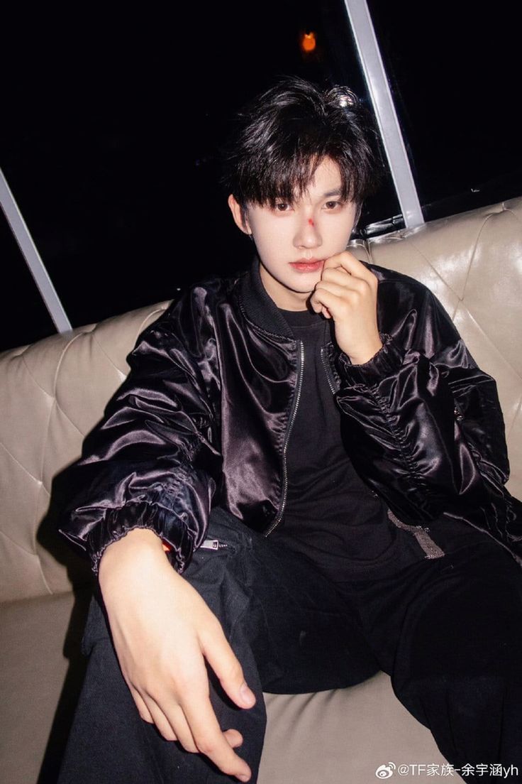 a young man sitting on top of a couch wearing a black shirt and pants with his hand in his mouth