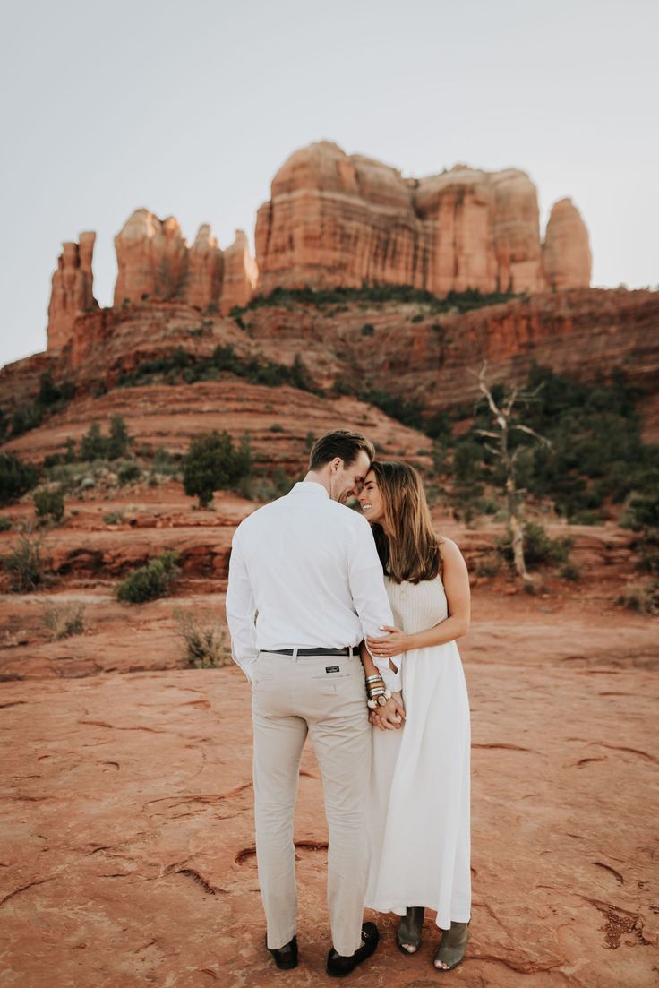 a man and woman are standing in front of some red rocks with the mountains behind them