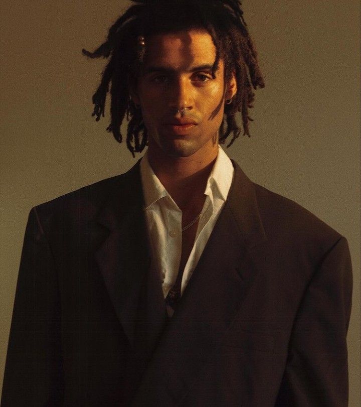 a man with dreadlocks standing in front of a white wall wearing a suit