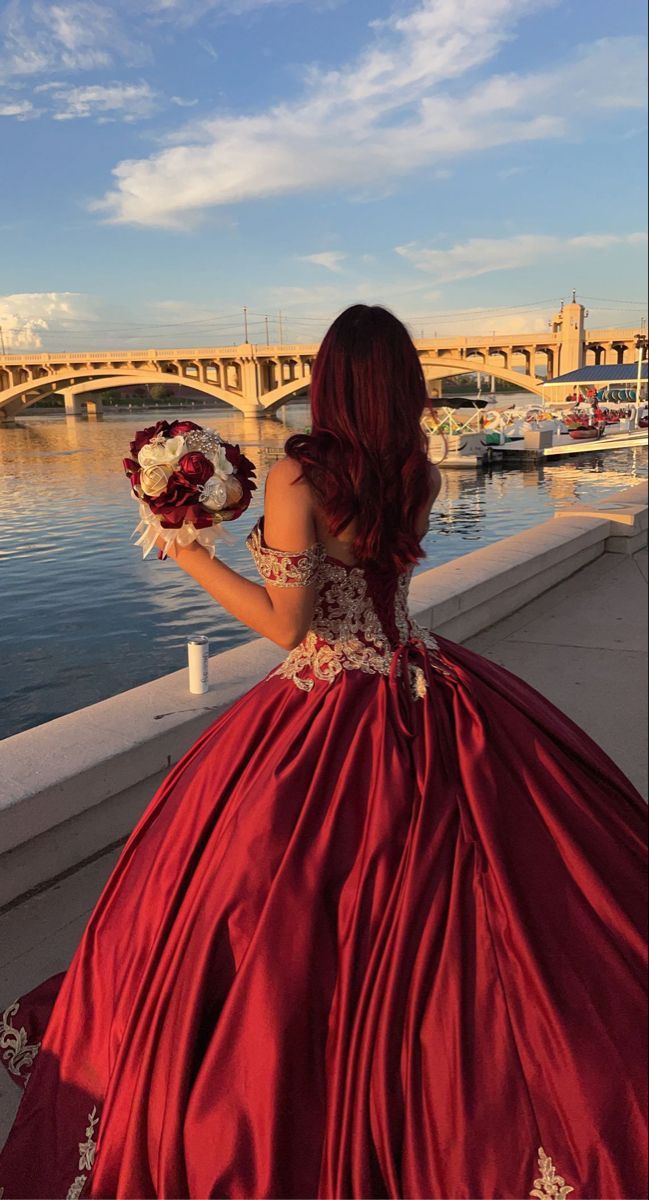 a woman in a long red dress is looking at the water and bridge that goes over it