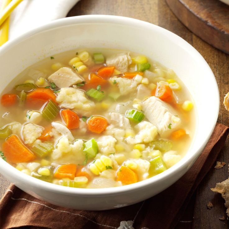 a white bowl filled with chicken and vegetable soup next to sliced bread on a wooden table