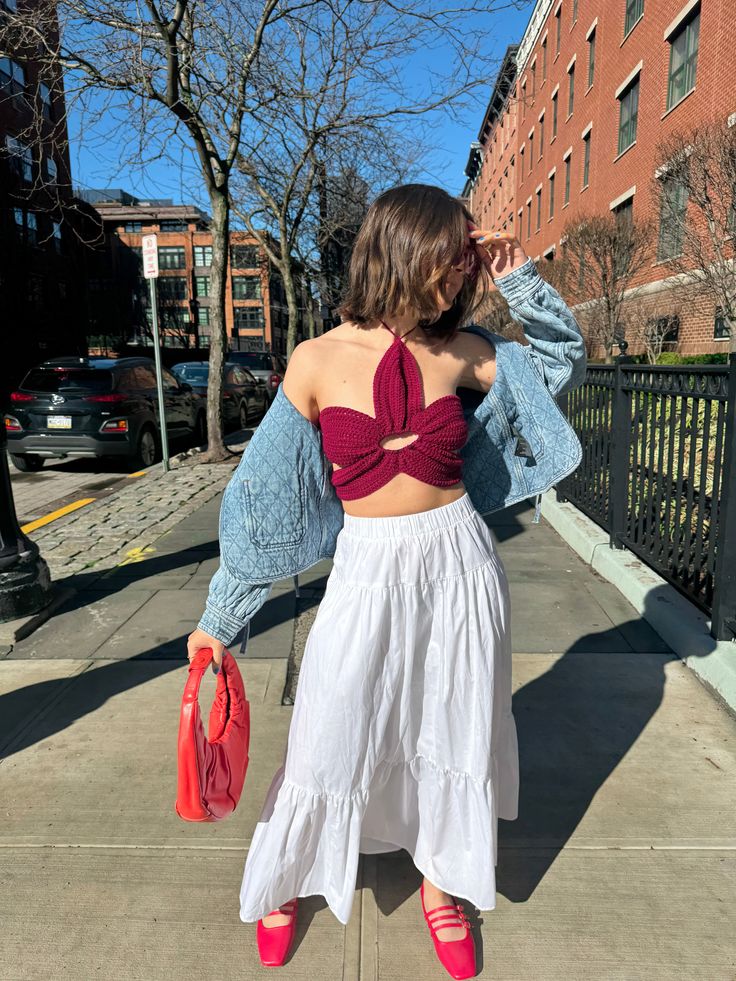 a woman in a red top and white skirt is standing on the sidewalk with her hands behind her head