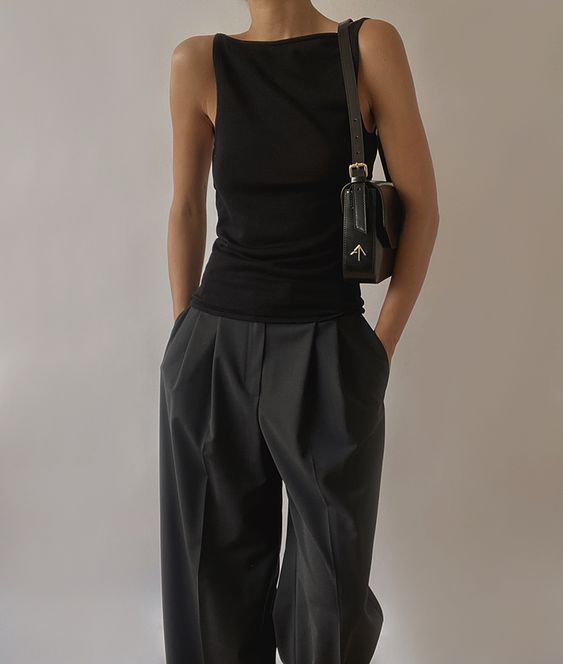 a woman standing in front of a white wall wearing black pants and a black bag