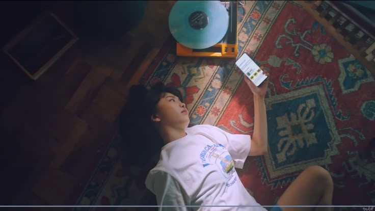 a woman laying on top of a rug next to a blue record player with an electronic device in her hand