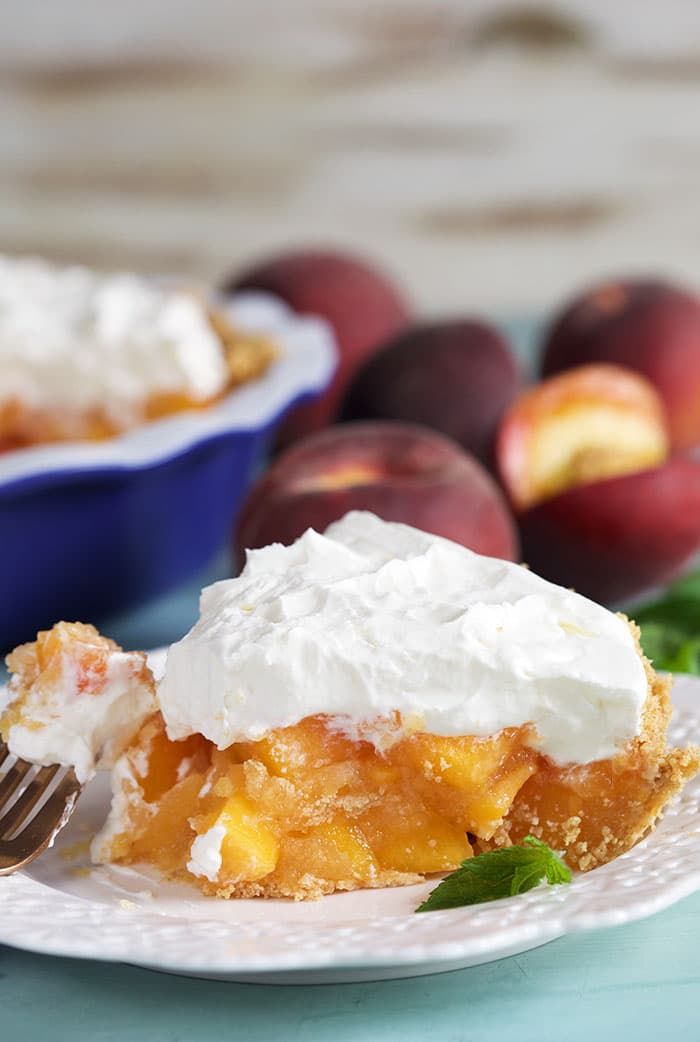 a piece of peach pie on a plate with a fork and bowl of fruit in the background