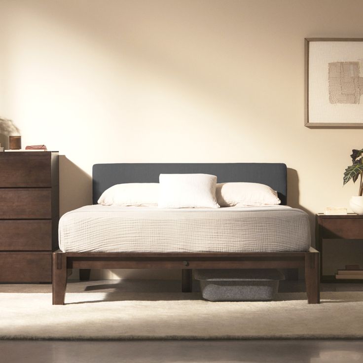 a bed with white sheets and pillows in a room next to a dresser, mirror and plant