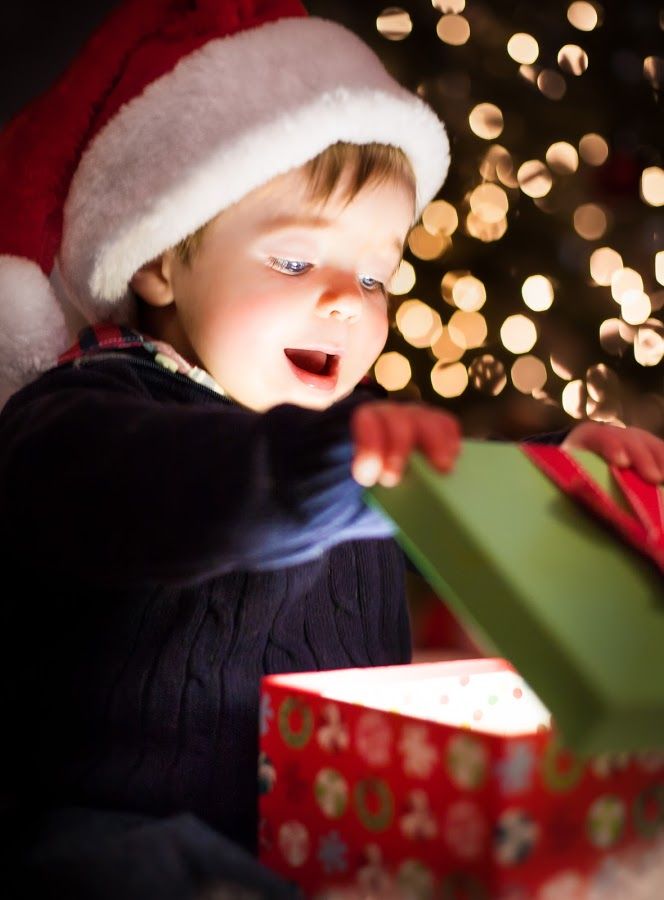 a small child in a santa hat opening a gift box with christmas lights behind it