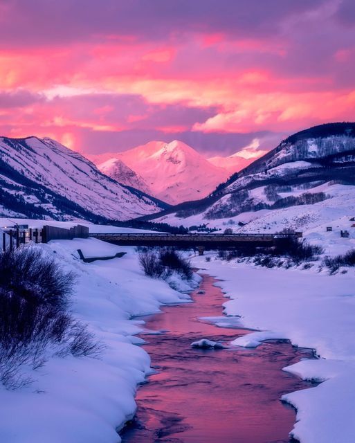 a river running through a snow covered field with mountains in the background and pink sky