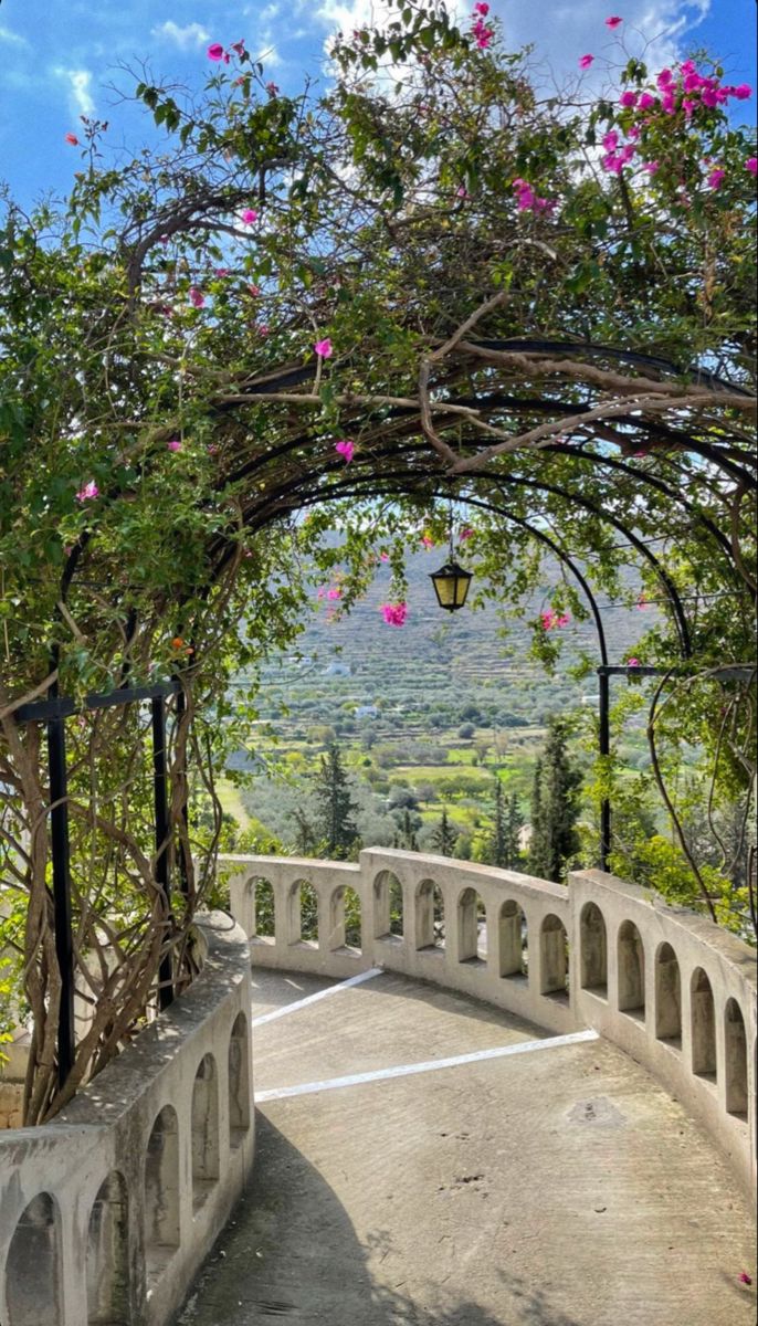 an arch with flowers growing over it on the side of a road that is surrounded by greenery