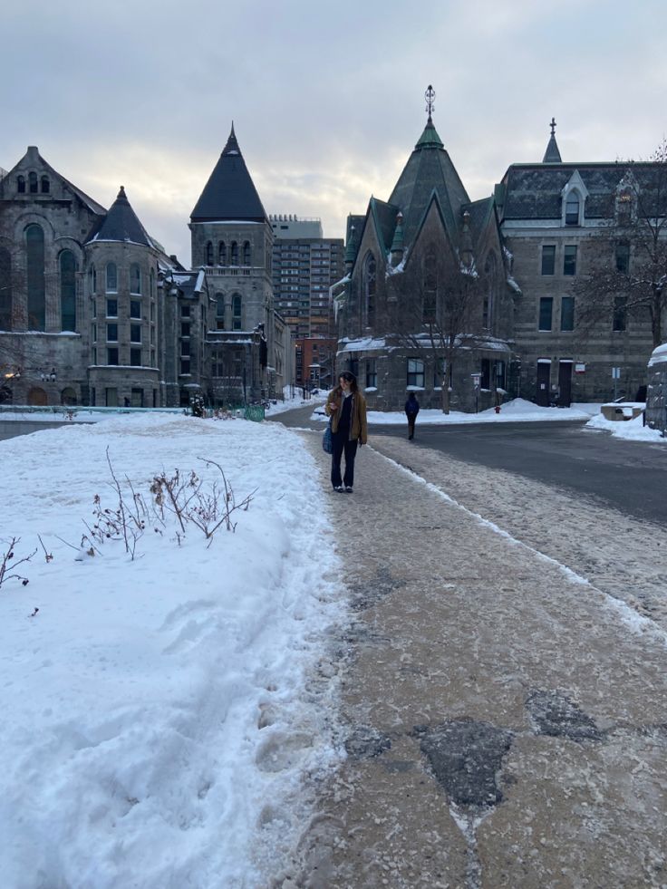 two people walking down a snow covered sidewalk in front of buildings and trees with no leaves on them