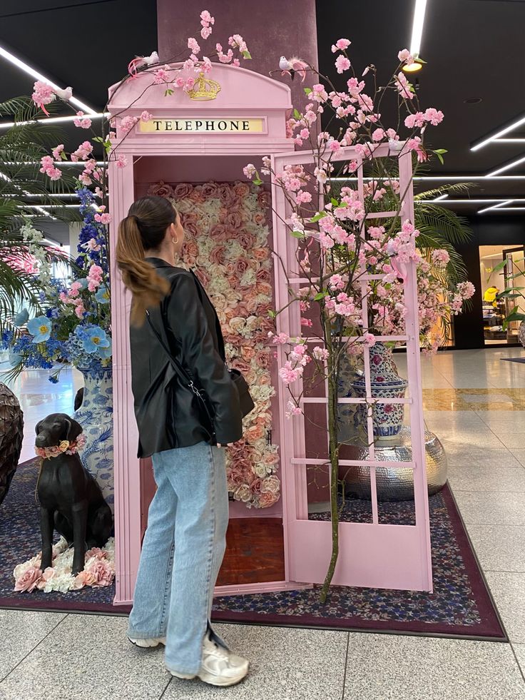 a woman standing in front of a pink phone booth with flowers on the wall and a dog sitting next to it