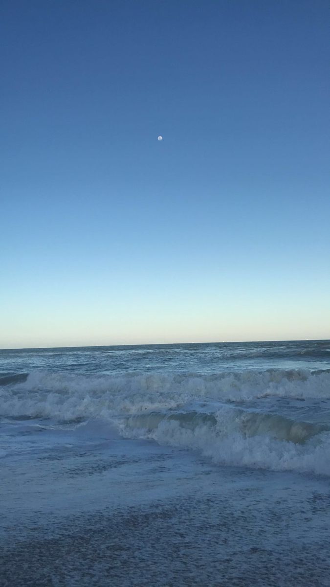 an ocean view with waves crashing on the shore and a half moon in the sky